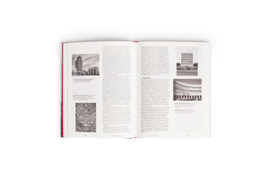 Draufsicht eines aufgeschlagenen Buches mit rotem Einband auf weißem Grund. Die Seiten zeigen deutschen Text über Architektur und Stadtplanung, illustriert mit mehreren Schwarz-Weiß-Fotografien von Gebäuden und einem Stadtplan.