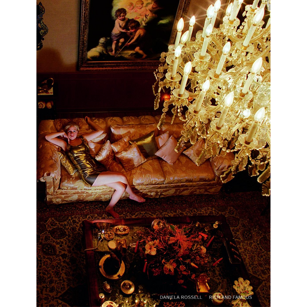 Eine Aufnahme aus der Vogelperspektive zeigt eine Frau in einem goldenen Kleid, die in einem opulenten Raum auf einem Sofa unter einem großen Kristallleuchter liegt.