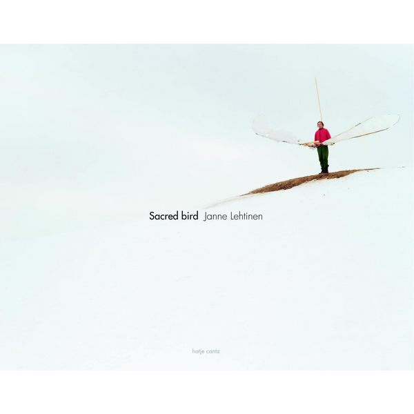 Das Buchcover von Sacred bird von Janne Lehtinen zeigt eine Person in einer roten Jacke, die auf einem schneebedeckten Hügel steht und ein Paar großer, durchsichtiger Flügel hält.