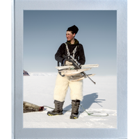 Ein junger Jäger in traditioneller Winterkleidung mit Fellhose und Gewehr steht in einer weiten Schneelandschaft, neben ihm liegt eine erlegte Robbe.