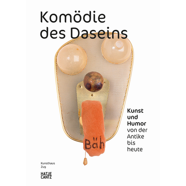Das Buchcover von Komödie des Daseins zeigt eine abstrakte Gesichtsskulptur aus goldenem Gitter. Zwei Glaskugeln dienen als Augen und eine orangefarbene Stoffzunge mit der Aufschrift Bäh hängt herab.