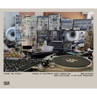 Ein Foto eines elektronischen Musikstudios, das mit modularen Synthesizern, unzähligen Kabeln, Lautsprechern und einem Plattenspieler im Vordergrund gefüllt ist.