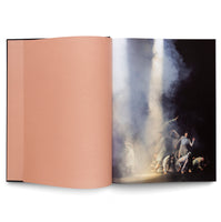 Ein aufgeschlagenes Buch mit einer leeren, lachsfarbenen linken Seite und einer Fotografie auf der rechten Seite, die eine Gruppe von Tänzern in ausdrucksstarken Posen auf einer dunklen, nebligen Bühne zeigt, die von einem hellen Lichtstrahl von oben beleuchtet wird.