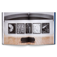 Ein aufgeschlagenes Buch, das eine Fotografie einer Galerieausstellung zeigt, in der fünf große, gerahmte Schwarz-Weiß-Bilder in einer Reihe an einer weißen Wand hängen, mit einer schwarzen Bank davor auf einem hellen Holzboden.