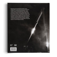Die schwarze Rückseite des Buches Robert Longo: The Acceleration of History. Links befindet sich ein Textblock mit Informationen zur Ausstellung, Logos und einem Barcode. Rechts ist eine dramatische Schwarz-Weiß-Kohlezeichnung zu sehen, die einen hellen Lichtstrahl zeigt, der diagonal durch eine dunkle, rauchige Szene verläuft.