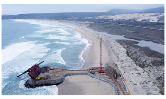 Luftaufnahme einer Küstenbaustelle an einem Sandstrand, wo ein großer roter Kran und Bagger auf einer Mole arbeiten, an deren Ende ein großes, rostiges Metallobjekt schräg im Wasser steckt, während sich im Hintergrund Dünen und Hügel erstrecken.