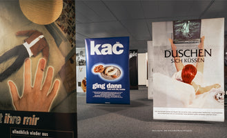 Eine Ausstellungshalle mit mehreren großen Postern auf einem grauen Teppichboden. Links ein Plakat mit einer leuchtenden Hand und einer kopflosen Figur auf einem Bett. In der Mitte ein blaues Plakat mit dem Wort kac und einer Abbildung von Weißwürsten. Rechts ein Plakat mit einer rothaarigen Person in einer Badewanne, die mit den Füßen einen Fernseher hält.
