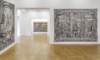 Eine Kunstgalerie mit Holzboden und weißen Wänden, an denen große, detailreiche Wandteppiche hängen. Rechts im Vordergrund ist ein schwarz-weißer Teppich mit den Teenage Mutant Ninja Turtles und einem Mann zu sehen, links ist ein Teil eines weiteren schwarz-weißen Teppichs mit einem futuristischen Motiv sichtbar. Durch einen Durchgang ist im Hintergrund ein weiterer Wandteppich zu sehen.