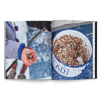 Ein aufgeschlagenes Buch mit einem Foto, das sich über zwei Seiten erstreckt. Links hält ein Kind in einem Wintermantel eine Handvoll Vogelfutter, die Fingernägel sind blau lackiert. Rechts steht ein großer weißer Eimer mit blauem Rand, gefüllt mit einer bunten Mischung aus Samen und Nüssen.