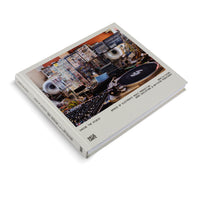 Ein cremefarbenes Buch mit dem Titel Inside the Studio liegt auf einer weißen Oberfläche. Das Cover zeigt ein Foto eines komplexen elektronischen Musikstudios mit vielen Synthesizern, Kabeln und einem Plattenspieler.