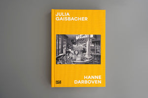Ein Buch mit einem leuchtend gelben Leineneinband liegt auf einer grauen Fläche. Auf dem Cover stehen in weißer Schrift die Namen Julia Gaisbacher und Hanne Darboven. Darunter ist ein Schwarz-Weiß-Foto eines vollgestellten Raumes mit einem Modell des Empire State Buildings abgebildet.
