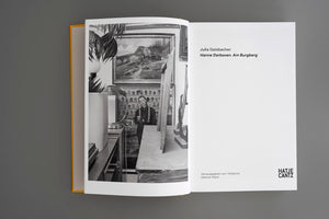 Ein aufgeschlagenes Kunstbuch mit gelbem Einband liegt auf einer grauen Oberfläche. Die linke Seite zeigt eine Schwarz-Weiß-Fotografie der Künstlerin Hanne Darboven in ihrem vollgestellten Atelier. Die rechte, weiße Seite listet den Buchtitel, die Autorin und den Verlag Hatje Cantz auf.