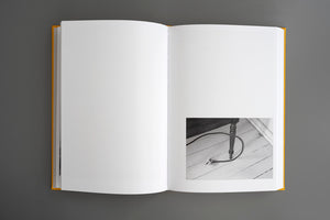 Ein aufgeschlagenes Buch mit gelbem Einband liegt auf einer grauen Fläche. Die linke Seite ist leer, während auf der rechten Seite unten ein kleines Schwarz-Weiß-Foto eines Netzsteckers und eines Kabels auf einem Holzboden neben einem Tischbein zu sehen ist.