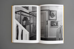 Ein aufgeschlagenes Buch mit gelbem Einband, das eine Schwarz-Weiß-Fotografie eines Treppenabsatzes zeigt. Links steht hinter einem weißen Geländer eine Glasvitrine mit einem hölzernen Pferdekopf. Rechts an der Wand hängen neben einer Tür zwei gerahmte Bilder.
