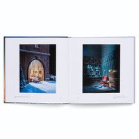Ein aufgeschlagenes Fotobuch liegt auf einer weißen Oberfläche. Die linke Seite zeigt eine verschneite Nachtszene mit Blick durch ein Bogenfenster in ein warm beleuchtetes Esszimmer. Die rechte Seite zeigt eine dunkle, holzgetäfelte Bibliothek mit einem roten Sessel und einem Bücherregal.