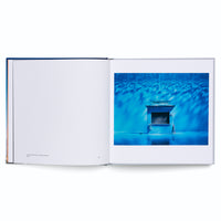 Ein aufgeschlagenes Fotobuch, das auf der rechten Seite eine Fotografie in leuchtenden Blautönen zeigt. Das Bild stellt eine unter Wasser stehende Trinkhalle vor einer gekachelten Wand dar, auf der sich Lichtreflexe bewegen.