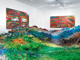 Eine Kunstinstallation in einer Galerie mit weißen Wänden. Im Vordergrund ist eine Landschaft aus Haufen bunter Farbpigmente in Grün, Blau, Gelb und Orange aufgeschüttet, durch die ein Pfad führt. Im Hintergrund hängen zwei große, farbenfrohe abstrakte Gemälde.