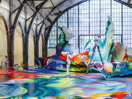 Zwei Künstler in Schutzanzügen arbeiten in einer großen Halle an einer riesigen, abstrakten und farbenfrohen Installation. Der gesamte Boden ist mit fließenden Farben bedeckt, und eine Person sprüht von einer Leiter aus Farbe auf eine große Skulptur, während die Säulen der Halle mit Plastikfolie abgedeckt sind.