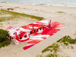 Luftaufnahme eines langen, verlassenen Gebäudes an einem Sandstrand, dessen Dach und umliegender Betonboden mit wirbelnder roter und weißer Farbe bedeckt sind, mit dem Meer im Hintergrund.