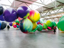 Eine große, helle Industriehalle ist mit riesigen, bunten, kugelförmigen Ballons gefüllt. Einige der Kugeln hängen, andere liegen auf dem Betonboden. Ein paar Menschen gehen zwischen den Ballons hindurch und wirken im Vergleich winzig.