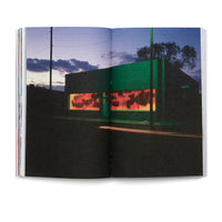 Ein aufgeschlagenes Buch, das eine Fotografie einer Straßenszene bei Dämmerung zeigt. Ein dunkelgrünes Gebäude steht an einer Ecke, seine Fenster leuchten in einem warmen Rotton, und darauf sind Graffiti-Schatten zu sehen. Auf der Straße im Vordergrund zeichnen sich die Lichtspuren eines Autos ab.