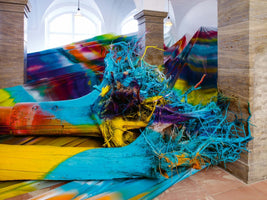 Eine große Kunstinstallation in einem Raum mit Steinsäulen, bestehend aus entwurzelten Baumstämmen, die mit leuchtenden Farben wie Türkis, Gelb und Orange besprüht sind. Das verworrene Wurzelwerk ist ebenfalls bunt gefärbt und im Hintergrund ist eine mehrfarbige Plane gespannt.