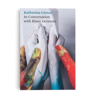 Buchcover von Katharina Grosse, In Conversation with Klaus Dermutz, das eine Nahaufnahme von farbverschmierten Händen zeigt, die einen weißen Handschuh mit einem Tuch reinigen.