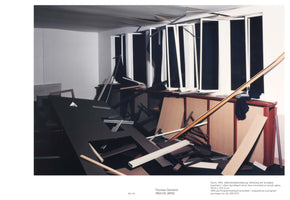 Fotografie eines zerstörten Büroraums, der als Modell aus Papier und Pappe nachgebaut wurde. Im Vordergrund ist ein umgestürzter schwarzer Tisch zu sehen, um den herum Trümmer verstreut sind. Eine lange Wand im Hintergrund hat Fenster mit kaputten, weißen Jalousien, die den Blick auf eine schwarze Fläche freigeben. Eine helle Holzleiste durchzieht das Bild diagonal.