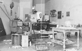 Schwarz-Weiß-Aufnahme eines vollgestellten Künstlerateliers. Eine Staffelei mit einem Gemälde steht im Raum, umgeben von Tischen, die mit Kunstzubehör, einem Globus und Farbdosen beladen sind. Auf dem Boden liegen Holzkisten, darunter eine von Canada Dry, sowie verstreute Papiere und Farbtuben. An den Wänden hängen verschiedene Kunstwerke und Regale.