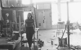 Schwarz-Weiß-Foto einer Frau, die in der Mitte eines großen, unordentlichen Künstlerateliers steht. Sie trägt ein gestreiftes Oberteil und dunkle Hosen. Der Raum ist gefüllt mit Leinwänden, einer Staffelei, einem Stuhl und einem Moped im hellen Hintergrund.