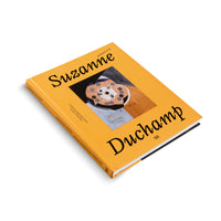 Ein gelbes Buch mit dem Titel Suzanne Duchamp Retrospective liegt schräg auf einem weißen Hintergrund. Auf dem Cover befindet sich die Abbildung eines abstrakten Gemäldes.