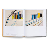 Ein aufgeschlagenes Kunstbuch zeigt zwei nebeneinander liegende Seiten mit farbenfrohen, modernistischen Gemälden von abstrakten Gebäuden, darunter befindet sich Text.