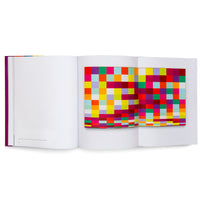 Ein aufgeschlagenes Buch mit lila Buchrücken auf weißem Hintergrund. Auf der rechten Seite ist ein ausklappbares, farbenfrohes Kunstwerk mit einem Mosaik aus bunten Quadraten zu sehen, während die linke Seite fast leer ist.