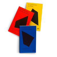 Drei Bücher mit leuchtend roten, blauen und gelben Einbänden liegen fächerförmig auf einer weißen Oberfläche. Jedes Buchcover zeigt eine große, schwarze, unregelmäßige geometrische Form und den Titel der Reihe On Architecture.