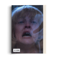 Die Rückseite eines Buches mit dem unscharfen Foto eines schreienden Gesichts mit blondem Haar, auf dem ein Handabdruck zu sehen ist. Unten links befindet sich ein Barcode und rechts der helle Stoffrücken des Buches.